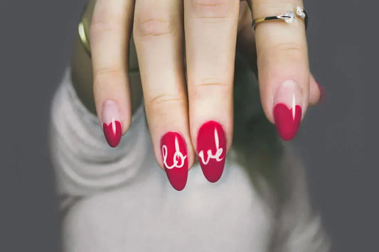 Close-up of a hand with almond-shaped nails featuring a red manicure. Two nails display heart designs, while the middle nails spell out 'love' in white cursive.