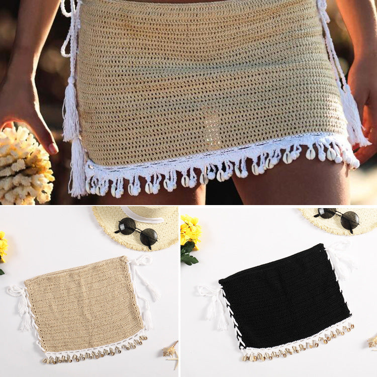 A person wearing a beige crochet skirt with white tassels, standing outdoors. Below are two crochet skirts laid flat, one beige with white tassels and one black with white tassels. The scene includes a straw hat and sunglasses.