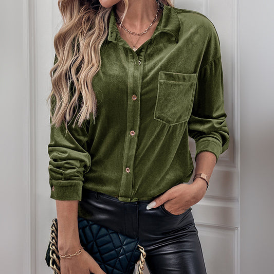 A person wearing a green corduroy shirt and black leather pants is standing indoors. The shirt is buttoned with a collar and chest pocket. They have long, wavy blond hair, and are accessorized with a gold necklace and belt.