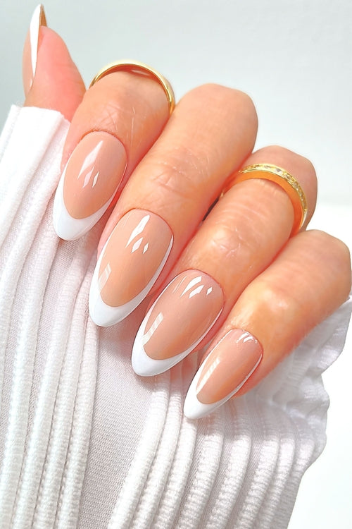 A hand with long, almond-shaped nails featuring a French manicure. The tips are white, and the base is a natural pink. The person is wearing gold rings and a white, pleated cuff on their wrist.