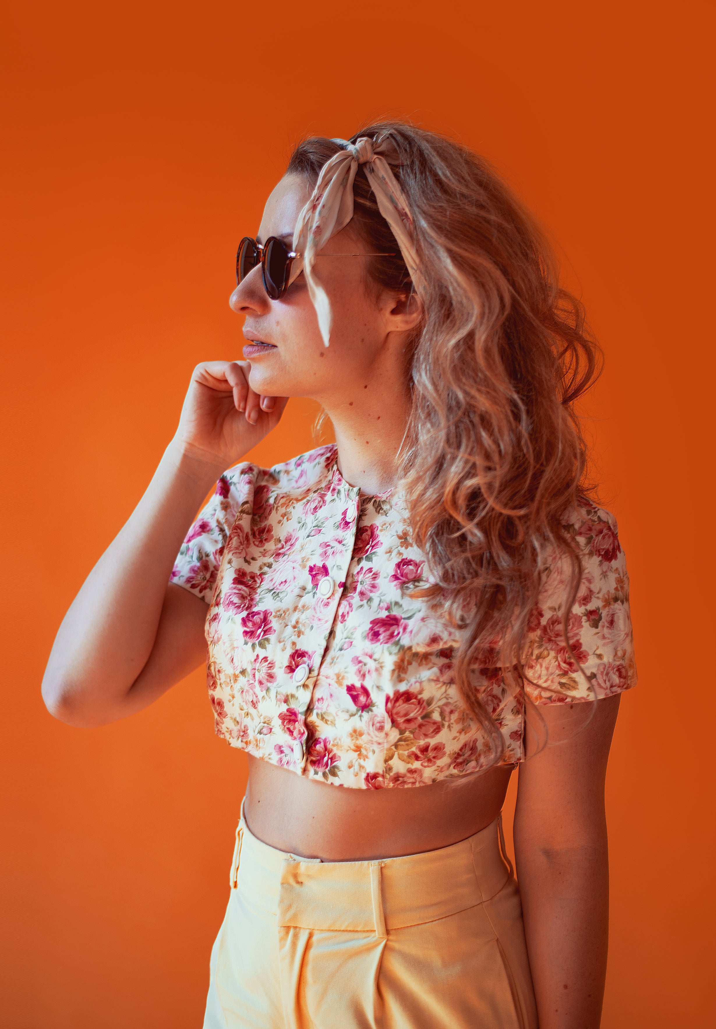 A woman with long hair wearing sunglasses, a floral crop top, and yellow pants stands against an orange background, gazing to the side.