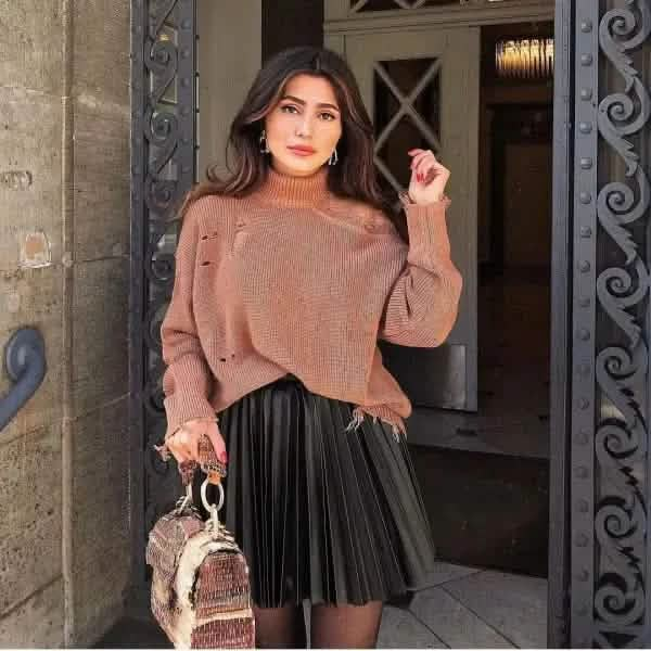 A woman wearing a peach turtleneck sweater and pleated black mini skirt.