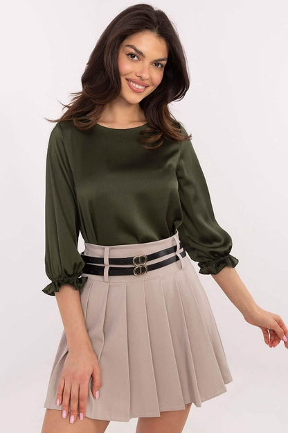 A woman wearing an olive green blouse and pleated beige skirt with a double-buckle belt.