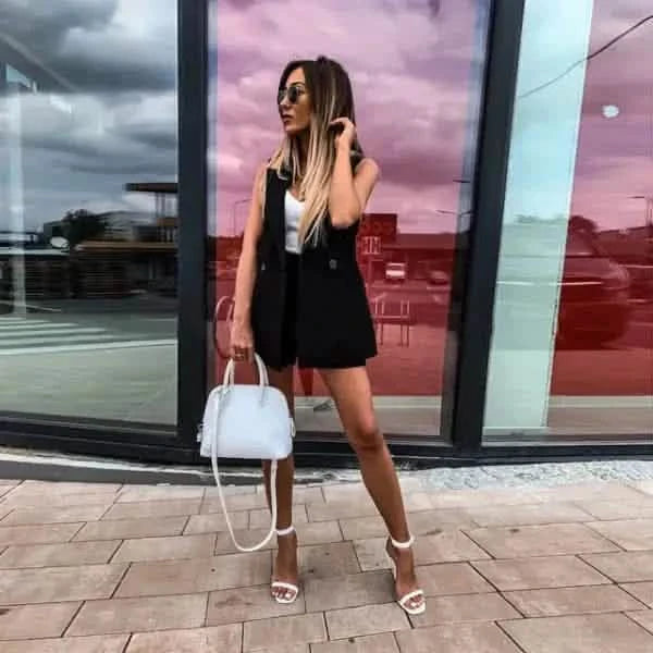 A person in a black sleeveless dress and white heels posing with a white handbag.